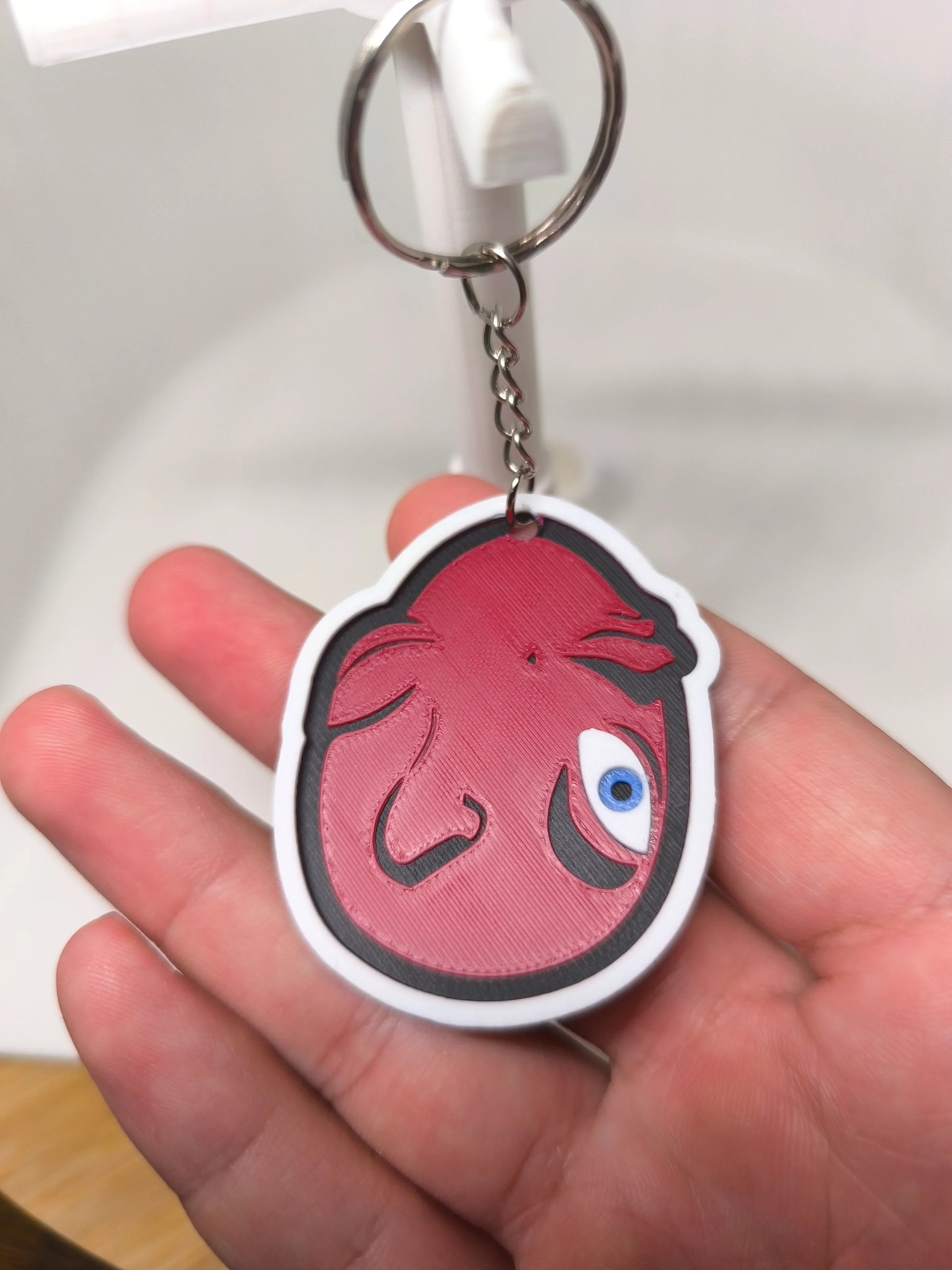 Porte-clés avec tête stylisée rouge et œil bleu tenu dans une main, fond clair