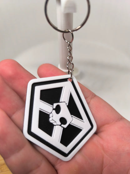 Porte-clés noir et blanc en forme de blason avec un crâne stylisé, tenu dans une main