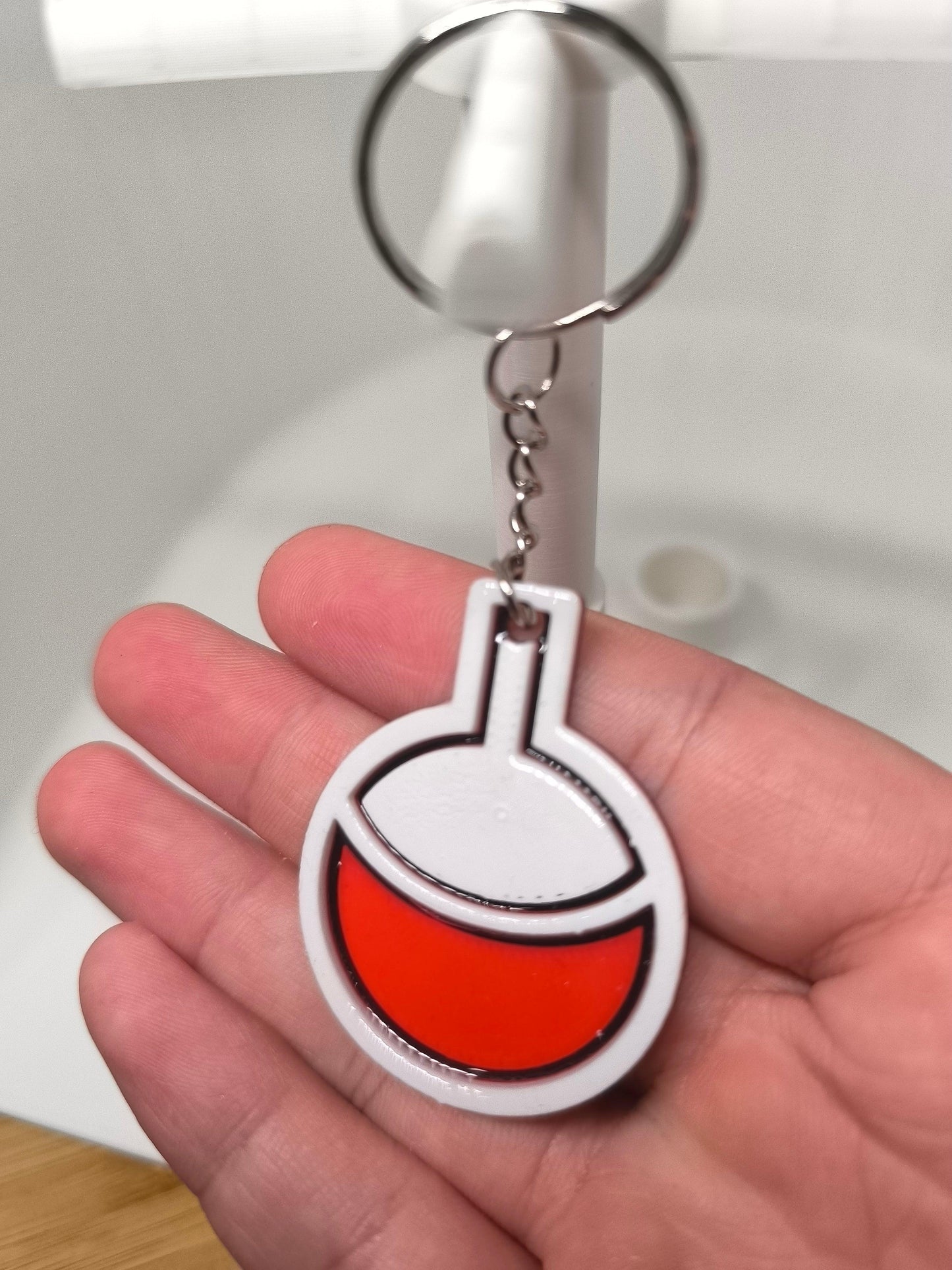 Porte-clés en forme de fiole de laboratoire avec liquide rouge tenu dans une main devant un lavabo blanc