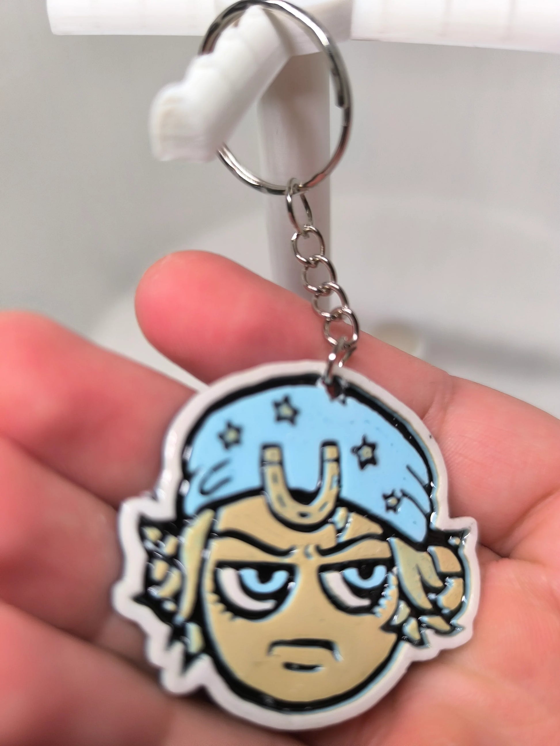 Porte-clés avec tête de personnage cartoon bleu et beige, tenu à la main, fond blanc flou