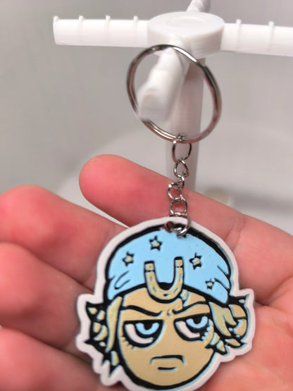 Porte-clés avec illustration d'un visage caricatural bleu et beige sur fond blanc, tenue à la main